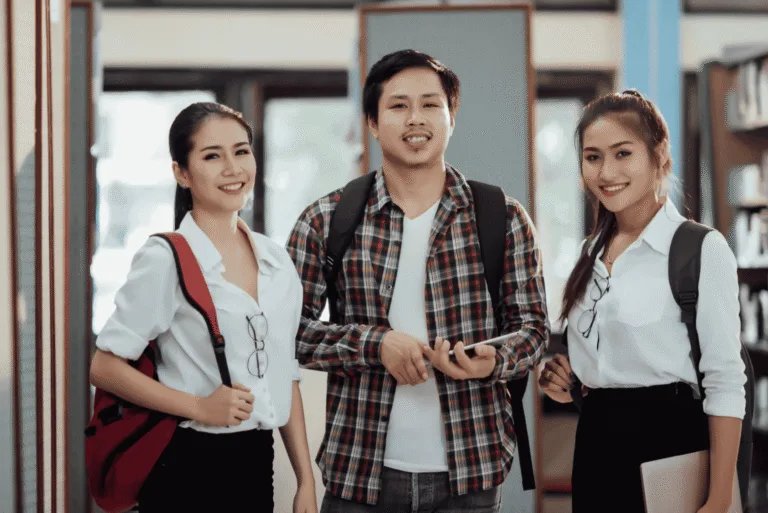Three students standing together in a library surrounded by bookshelves.